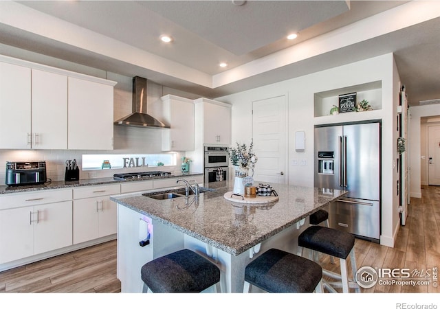 kitchen with an island with sink, appliances with stainless steel finishes, light wood-type flooring, wall chimney exhaust hood, and sink