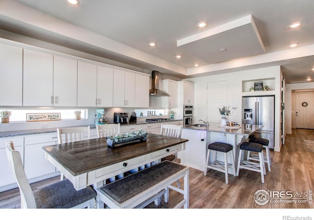 kitchen with a kitchen island with sink, wall chimney exhaust hood, stainless steel appliances, white cabinetry, and light hardwood / wood-style floors