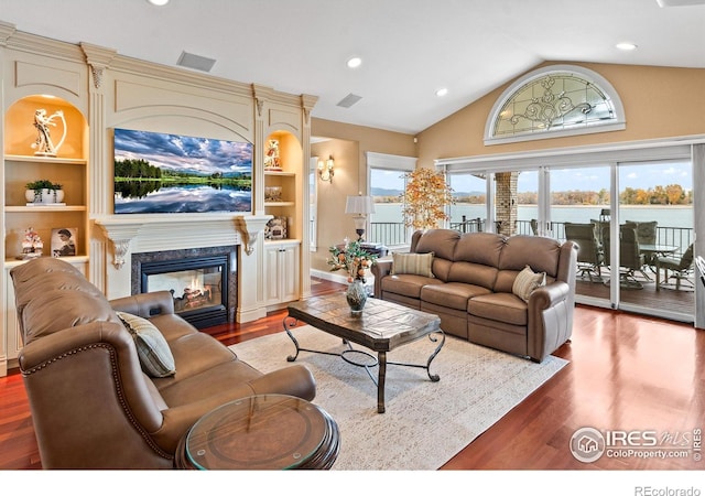 living room with hardwood / wood-style flooring, a premium fireplace, vaulted ceiling, built in features, and a water view