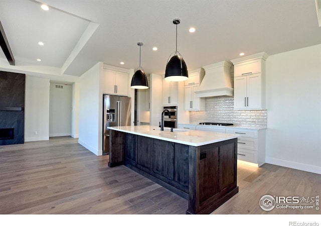 kitchen with appliances with stainless steel finishes, light wood-type flooring, premium range hood, and a kitchen island with sink