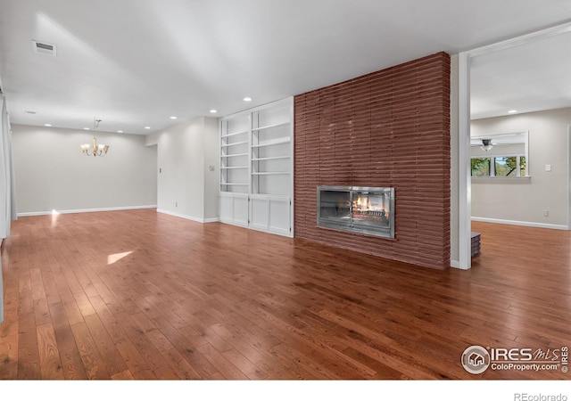 unfurnished living room with a brick fireplace, ceiling fan with notable chandelier, wood-type flooring, and built in features