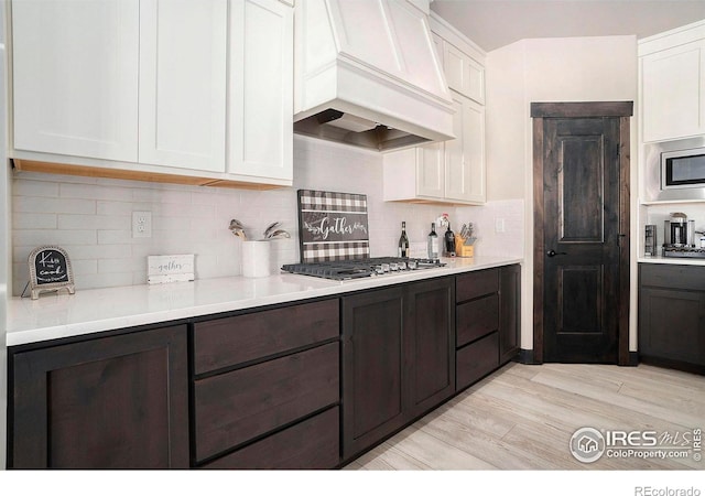 kitchen with appliances with stainless steel finishes, white cabinetry, dark brown cabinets, custom exhaust hood, and light wood-type flooring