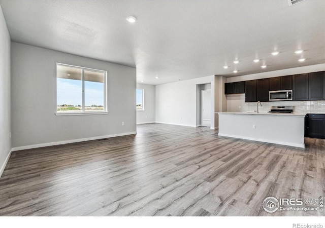 kitchen featuring an island with sink, backsplash, stainless steel appliances, dark brown cabinetry, and light hardwood / wood-style flooring