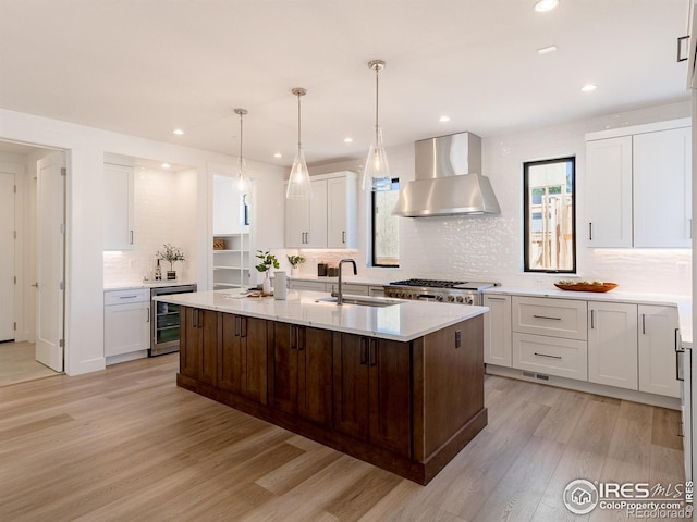 kitchen featuring wall chimney range hood, sink, light hardwood / wood-style floors, beverage cooler, and a center island with sink