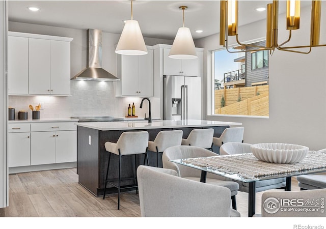 kitchen featuring wall chimney range hood, light wood-type flooring, hanging light fixtures, stainless steel appliances, and white cabinets