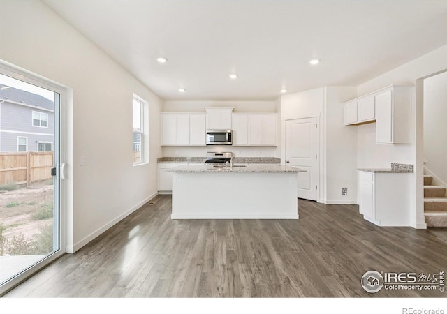 kitchen with appliances with stainless steel finishes, an island with sink, hardwood / wood-style floors, white cabinetry, and light stone counters