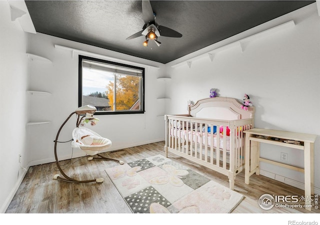 bedroom with ceiling fan, a textured ceiling, hardwood / wood-style floors, and a nursery area