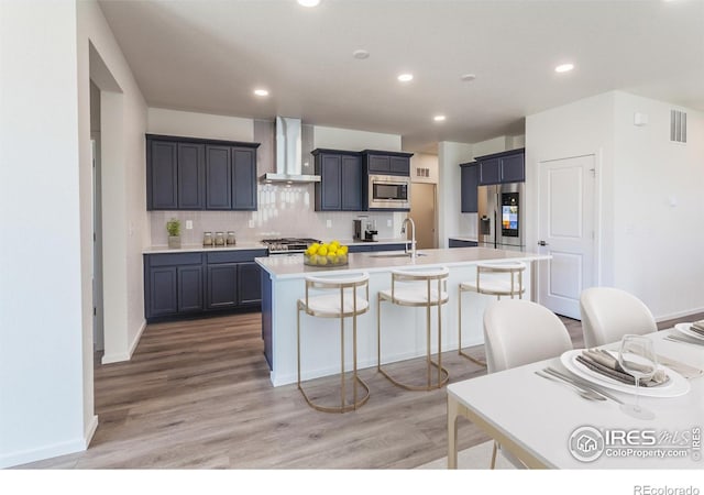 kitchen with wall chimney exhaust hood, a kitchen island with sink, stainless steel appliances, and wood-type flooring