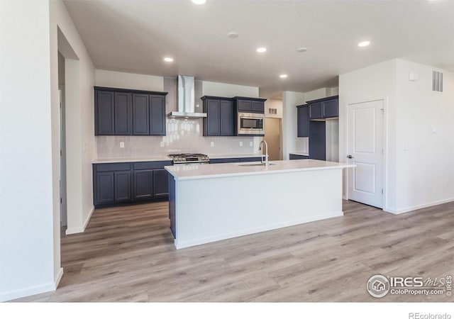 kitchen with stainless steel microwave, wall chimney exhaust hood, sink, and an island with sink