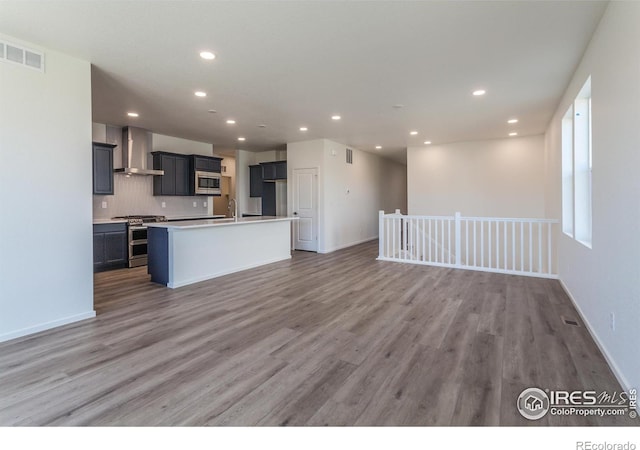 kitchen with wall chimney range hood, sink, appliances with stainless steel finishes, light hardwood / wood-style floors, and a center island with sink