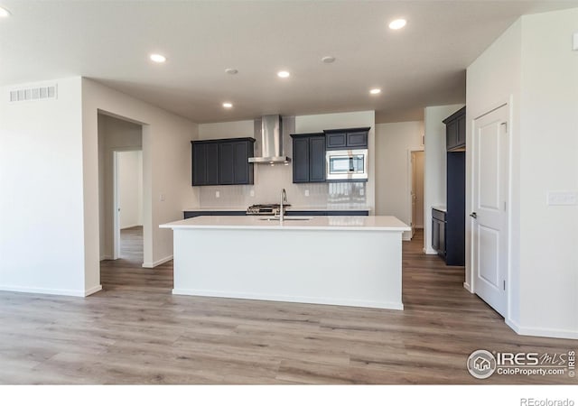 kitchen featuring sink, a kitchen island with sink, stainless steel microwave, and wall chimney exhaust hood