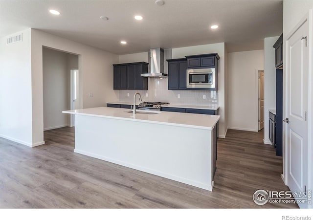 kitchen with sink, stainless steel microwave, an island with sink, decorative backsplash, and wall chimney exhaust hood