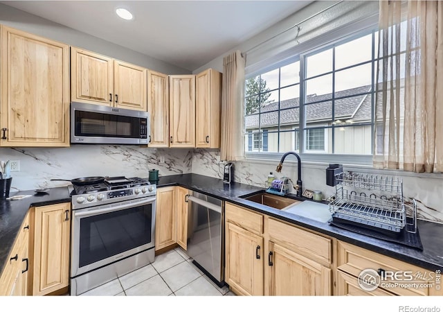 kitchen featuring light brown cabinetry, stainless steel appliances, dark countertops, a sink, and tasteful backsplash