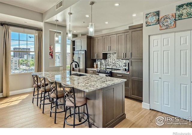 kitchen featuring light wood-type flooring, an island with sink, hanging light fixtures, stainless steel appliances, and light stone counters