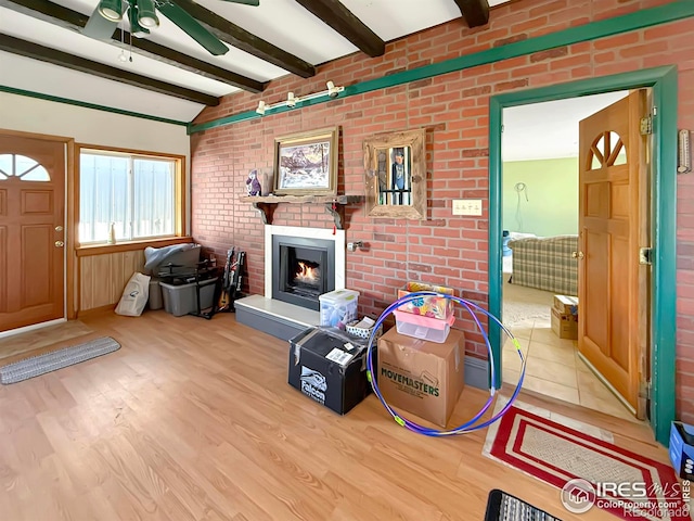 living area with a ceiling fan, brick wall, vaulted ceiling with beams, a lit fireplace, and light wood-style floors