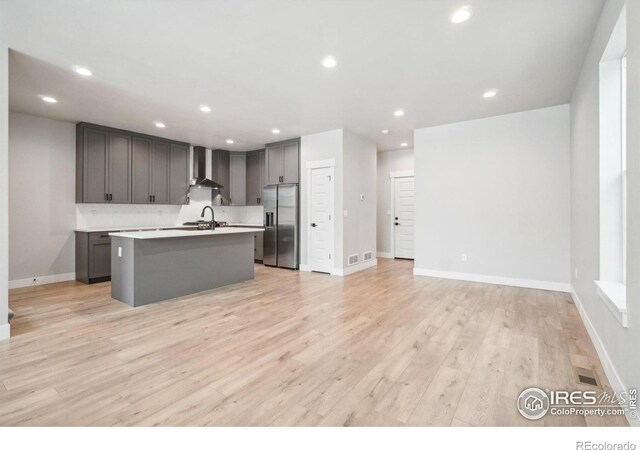 kitchen featuring sink, an island with sink, stainless steel fridge with ice dispenser, wall chimney range hood, and light wood-type flooring