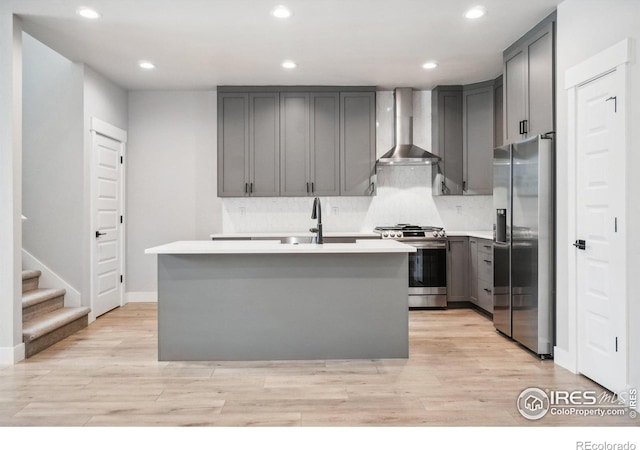 kitchen featuring light hardwood / wood-style flooring, an island with sink, gray cabinets, wall chimney range hood, and appliances with stainless steel finishes