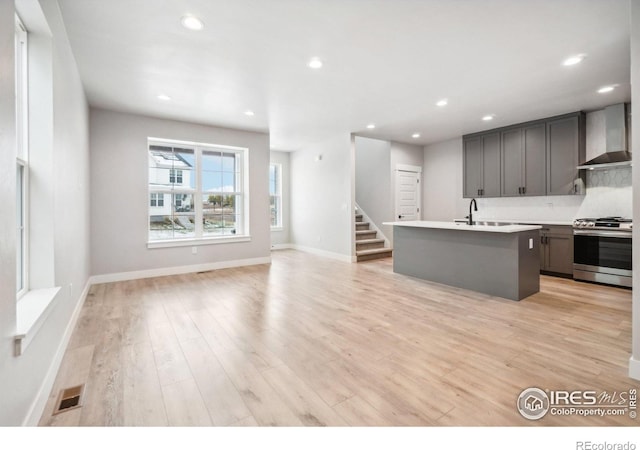 kitchen with light hardwood / wood-style floors, an island with sink, wall chimney range hood, gray cabinetry, and stainless steel gas stove