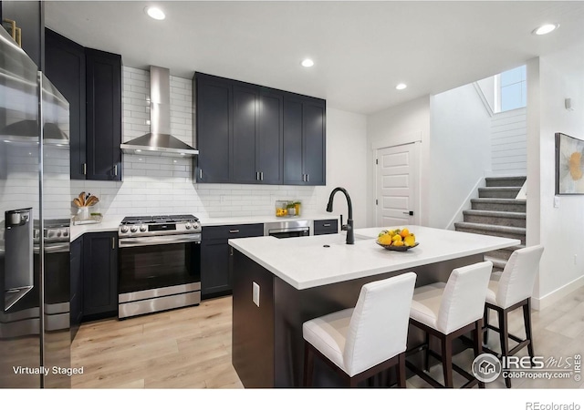 kitchen with wall chimney range hood, appliances with stainless steel finishes, an island with sink, and light hardwood / wood-style flooring