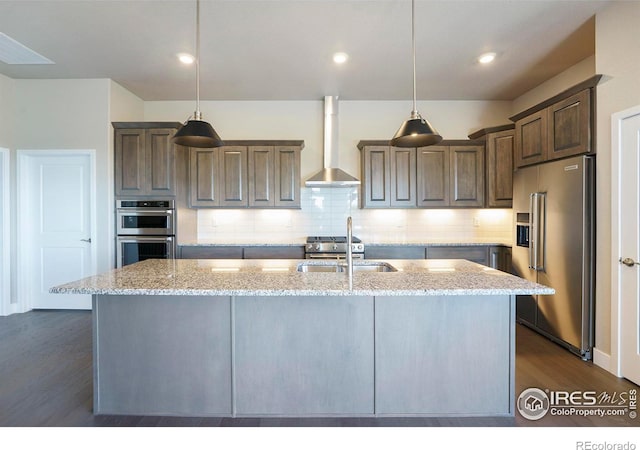 kitchen with dark wood-type flooring, wall chimney range hood, hanging light fixtures, light stone countertops, and appliances with stainless steel finishes