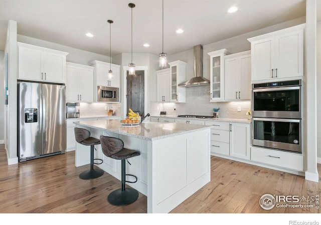 kitchen with an island with sink, wall chimney exhaust hood, light hardwood / wood-style floors, and stainless steel appliances