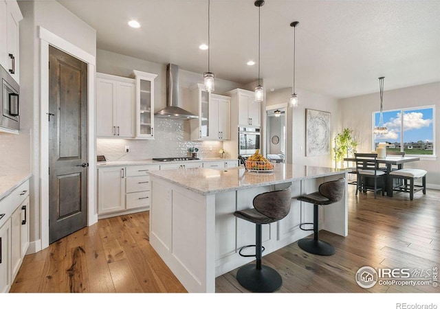 kitchen with wood-type flooring, stainless steel appliances, white cabinetry, wall chimney range hood, and a center island