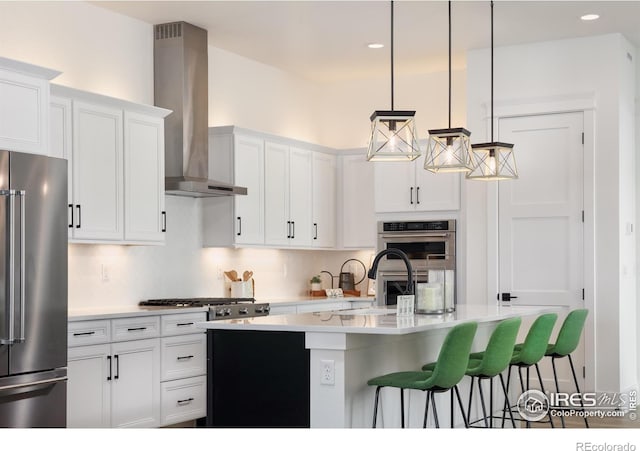 kitchen featuring a kitchen bar, appliances with stainless steel finishes, pendant lighting, wall chimney range hood, and a kitchen island with sink