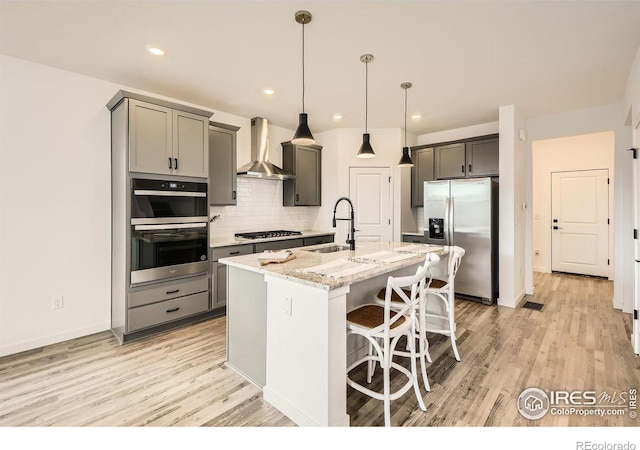 kitchen with wall chimney range hood, sink, light stone countertops, an island with sink, and appliances with stainless steel finishes