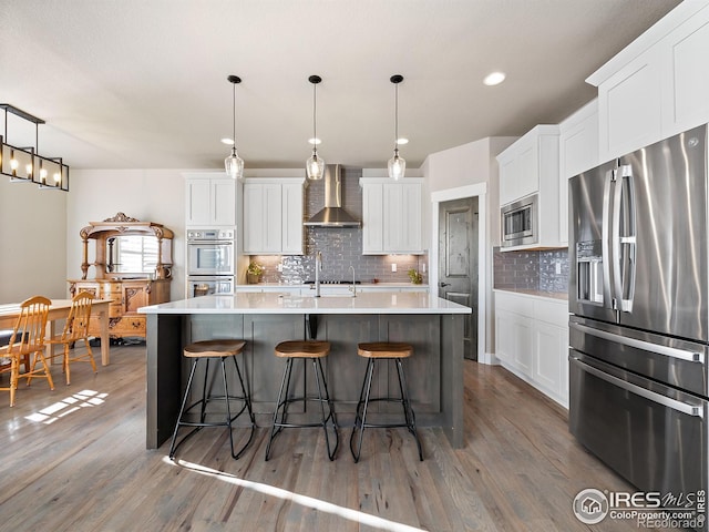 kitchen with stainless steel appliances, wood finished floors, a sink, wall chimney exhaust hood, and an island with sink