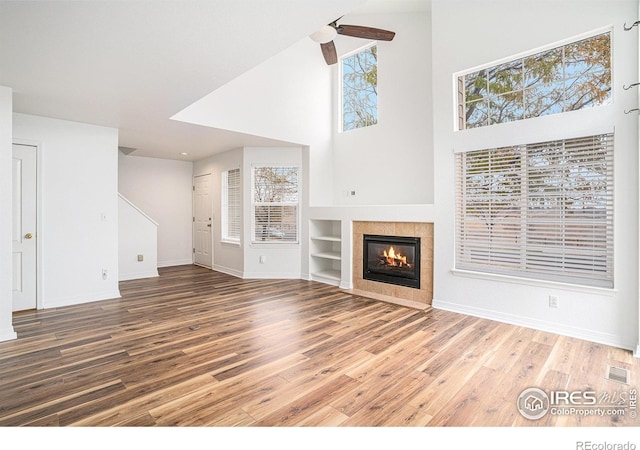 unfurnished living room with ceiling fan, a fireplace, a towering ceiling, wood-type flooring, and built in features