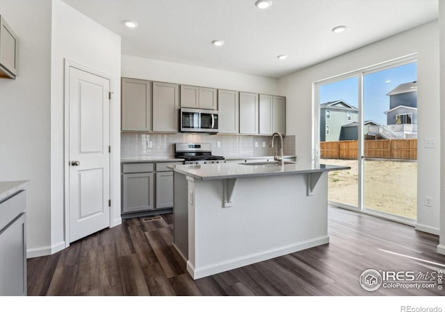 kitchen featuring stainless steel appliances, sink, backsplash, an island with sink, and dark wood-type flooring