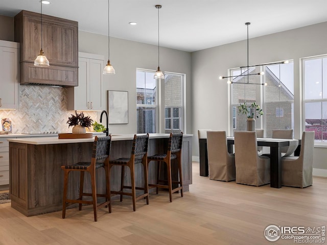 kitchen with a center island with sink, light wood-style flooring, light countertops, pendant lighting, and backsplash