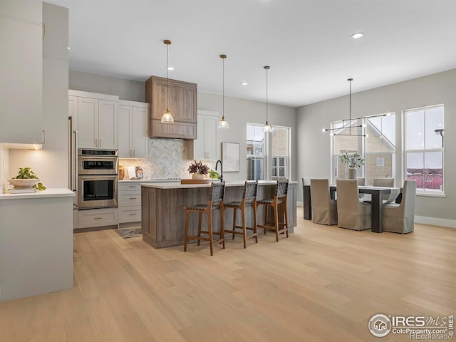 kitchen with decorative light fixtures, tasteful backsplash, light wood-type flooring, an island with sink, and stainless steel double oven