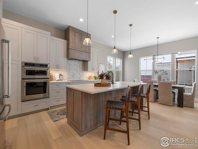 kitchen featuring an island with sink, appliances with stainless steel finishes, decorative backsplash, decorative light fixtures, and light wood-type flooring