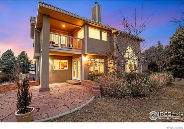 back of house at dusk featuring a patio, a chimney, stucco siding, fence, and a balcony