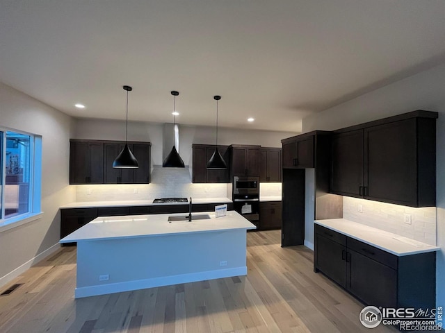 kitchen featuring wall chimney range hood, sink, an island with sink, tasteful backsplash, and light hardwood / wood-style floors