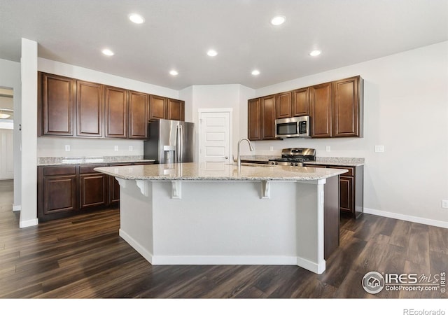 kitchen featuring appliances with stainless steel finishes, dark wood-type flooring, a sink, an island with sink, and a kitchen bar