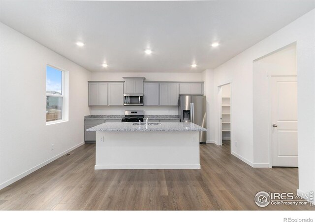 kitchen featuring appliances with stainless steel finishes, light stone counters, light hardwood / wood-style floors, gray cabinets, and an island with sink