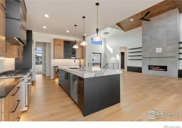 kitchen featuring wall chimney exhaust hood, pendant lighting, a center island with sink, appliances with stainless steel finishes, and light wood-type flooring