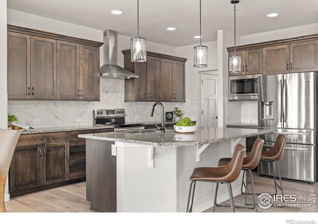 kitchen with wall chimney range hood, light stone counters, decorative light fixtures, a center island with sink, and appliances with stainless steel finishes