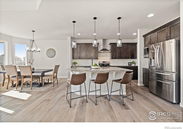 kitchen with wall chimney range hood, backsplash, pendant lighting, dark brown cabinets, and appliances with stainless steel finishes