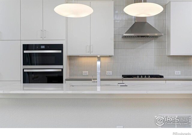 kitchen with stainless steel double oven, wall chimney range hood, light stone counters, black gas stovetop, and white cabinets