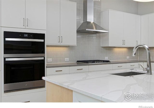 kitchen with light stone countertops, stainless steel appliances, sink, wall chimney range hood, and white cabinets