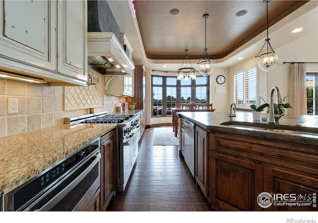 kitchen with appliances with stainless steel finishes, a raised ceiling, pendant lighting, dark stone countertops, and dark hardwood / wood-style floors