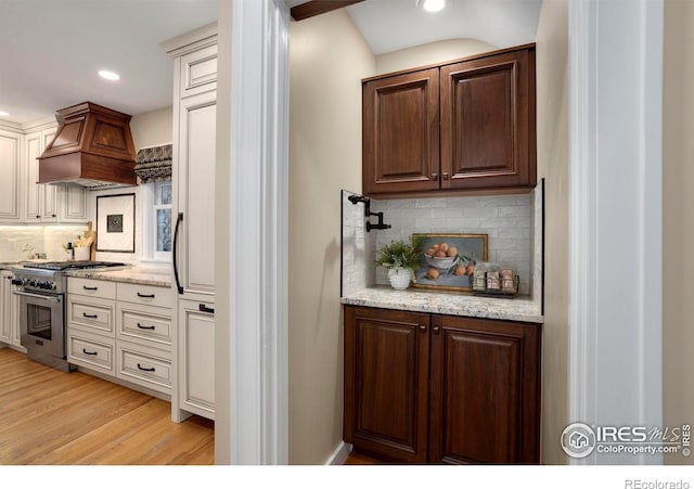 kitchen featuring custom exhaust hood, high end stainless steel range, light stone countertops, tasteful backsplash, and light hardwood / wood-style floors