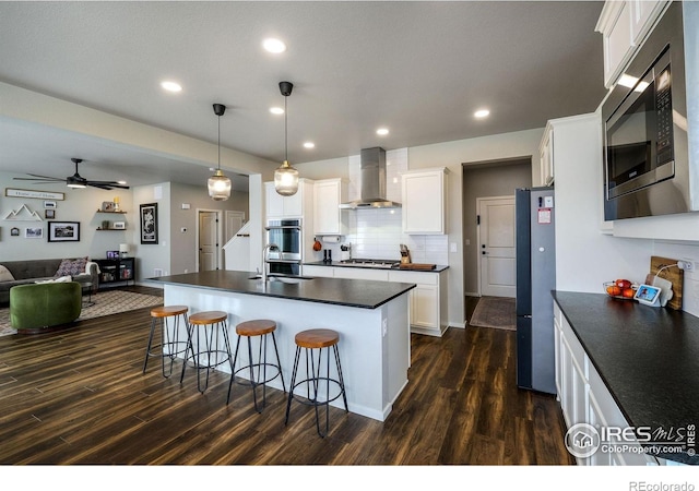 kitchen with stainless steel appliances, a kitchen island with sink, wall chimney range hood, dark hardwood / wood-style floors, and white cabinetry