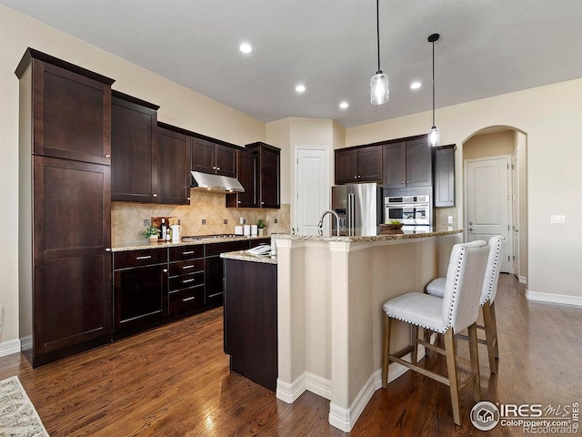 kitchen with a kitchen island with sink, hanging light fixtures, dark hardwood / wood-style floors, decorative backsplash, and appliances with stainless steel finishes