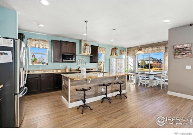 kitchen with pendant lighting, stainless steel refrigerator, built in microwave, a kitchen island with sink, and light stone countertops