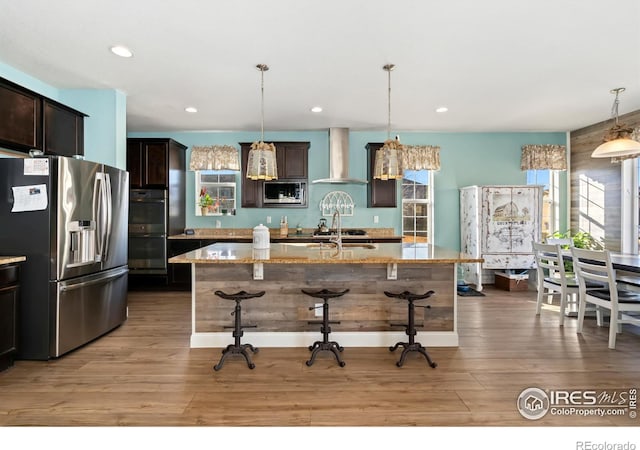 kitchen featuring an island with sink, hanging light fixtures, light stone counters, stainless steel appliances, and wall chimney range hood