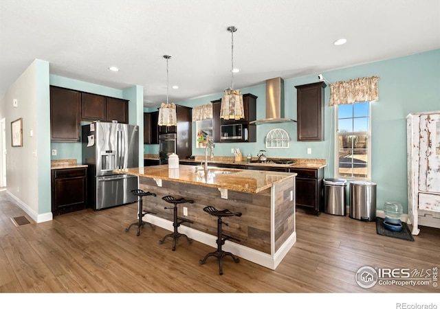 kitchen featuring wall chimney range hood, sink, appliances with stainless steel finishes, a center island with sink, and decorative light fixtures